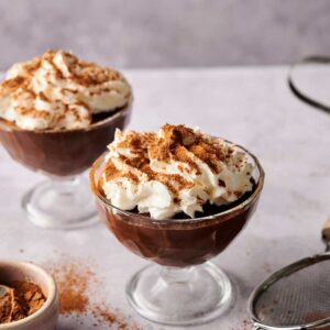 Two glass dessert cups filled with rich chocolate pudding, topped with whipped cream and a dusting of cocoa powder, sit on a light surface with cocoa and a sifter nearby.