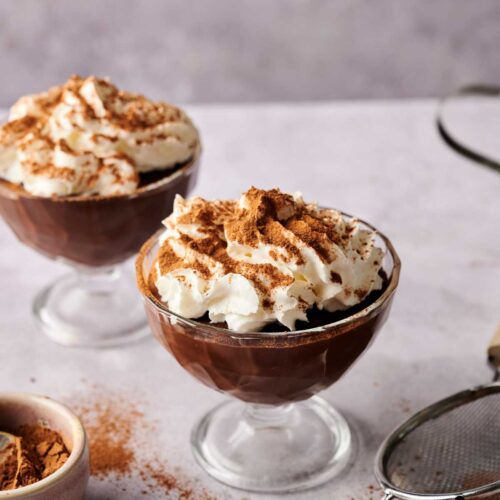 Two glass dessert cups filled with rich chocolate pudding, topped with whipped cream and a dusting of cocoa powder, sit on a light surface with cocoa and a sifter nearby.