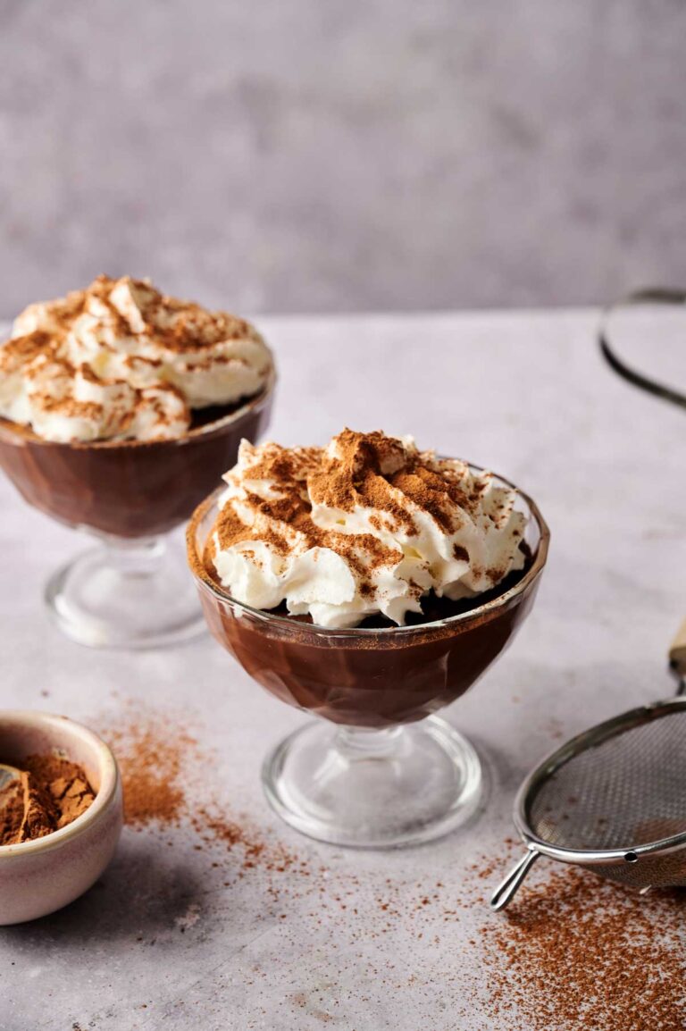 Two glass dessert cups filled with rich chocolate pudding, topped with whipped cream and a dusting of cocoa powder, sit on a light surface with cocoa and a sifter nearby.
