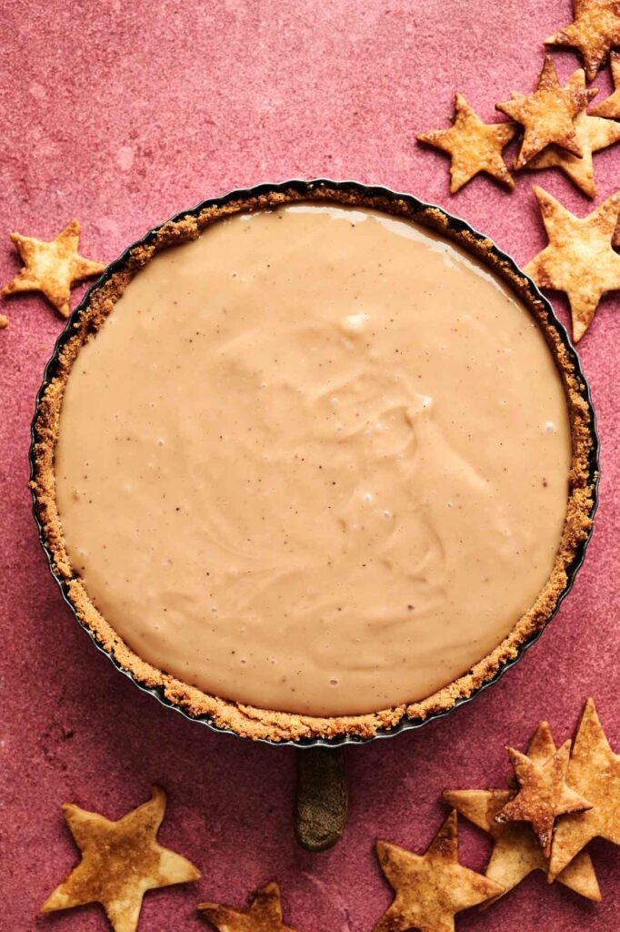 An Eggnog Pie with a smooth, light brown filling sits in a pan on a pink surface, surrounded by several star-shaped pastry pieces.