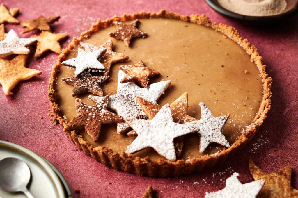 An Eggnog Pie with a smooth brown filling is topped with star-shaped cookies, some dusted with powdered sugar, on a textured pink surface.