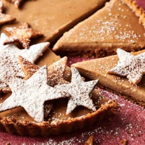 An Eggnog Pie with a crumb crust is sliced, topped with star-shaped cookies dusted with powdered sugar, on a pink surface.
