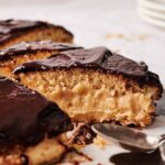A close-up of a sliced Boston Cream Pie, its creamy layer nestled between soft sponge cake and topped with glossy chocolate icing; a spoon rests nearby.