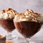 Two glass dessert cups filled with decadent chocolate mousse and chocolate pudding, topped with whipped cream and a sprinkle of cocoa powder, are placed on a light surface.