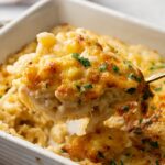 A close-up of a serving of Four Cheese Mac and Cheese being lifted from a casserole dish, topped with melted cheese and garnished with herbs.