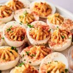 A plate of Million Dollar Deviled Eggs garnished with paprika and chopped parsley, arranged in rows on a white platter.