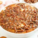 A round dish filled with a pecan praline topping over a creamy Pecan Pie Dip, with vanilla wafers and a bowl of pecans nearby.