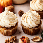 Three pumpkin cupcakes with swirled white frosting and a sprinkle of cinnamon, surrounded by autumn decorations including nuts, gourds, and dried plants.