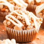 A close-up of Pumpkin Spice Muffins with Cream Cheese Frosting, topped with crumbly streusel and drizzled with white icing, resting on a wooden surface.
