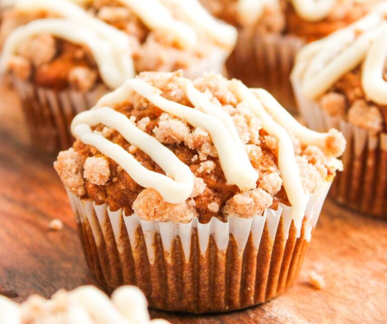 A close-up of Pumpkin Spice Muffins with Cream Cheese Frosting, topped with crumbly streusel and drizzled with white icing, resting on a wooden surface.