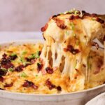 A Twice-Baked Potato Casserole topped with melted cheese, bacon bits, and chopped green onions, with a serving being lifted to reveal the irresistible stretchy cheese.