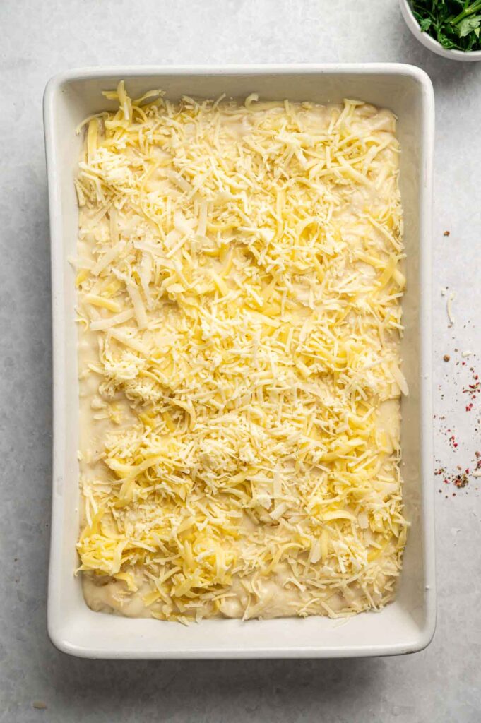 A rectangular baking dish filled with unbaked Four Cheese Mac and Cheese casserole, topped generously with shredded cheese, shown from above on a light surface.