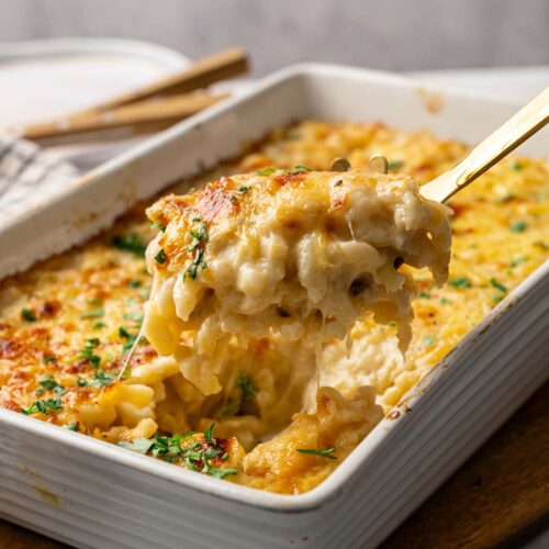 A close-up of Four Cheese Mac and Cheese being scooped from a white rectangular casserole dish, with melted cheese and parsley on top.