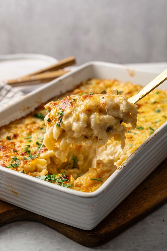 A close-up of Four Cheese Mac and Cheese being scooped from a white rectangular casserole dish, with melted cheese and parsley on top.