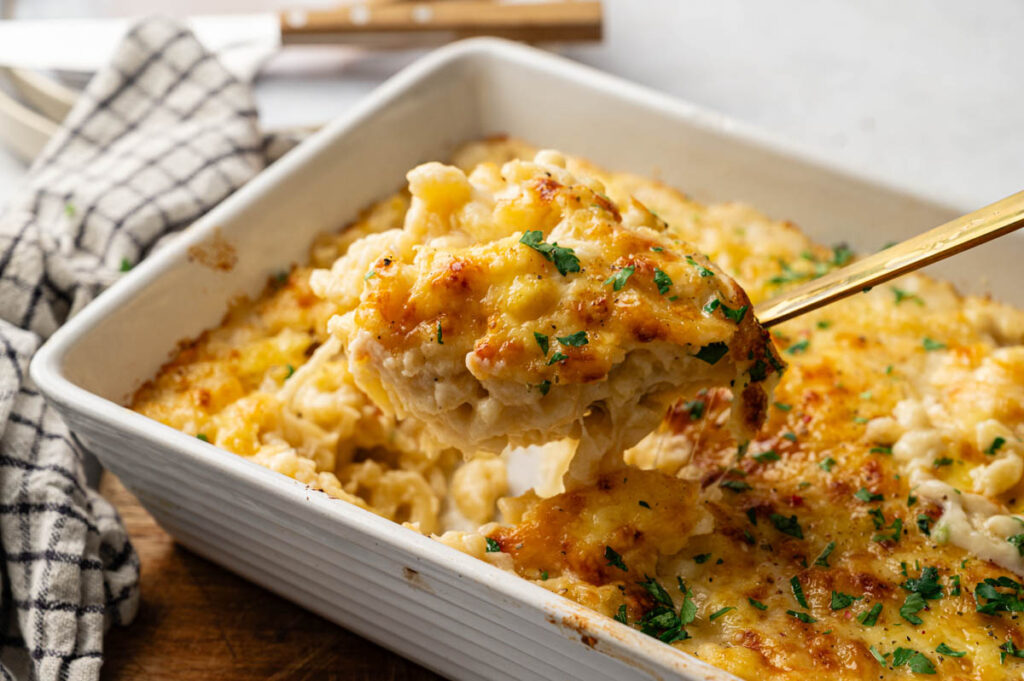 A close-up of Four Cheese Mac and Cheese in a white casserole dish, with a serving spoon lifting a portion. The dish is topped with melted cheese and sprinkled with chopped herbs.