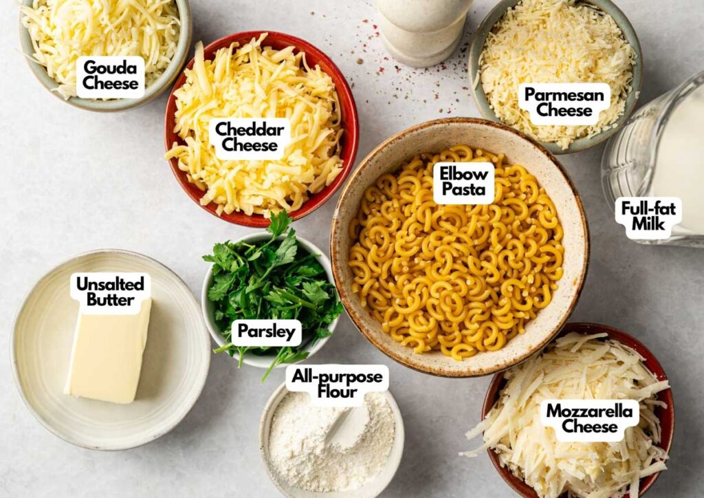 Bowls containing shredded gouda, cheddar, parmesan, mozzarella, elbow pasta, parsley, flour, butter, and a glass of full-fat milk are arranged on a white surface&mdash;perfect for making Four Cheese Mac and Cheese.