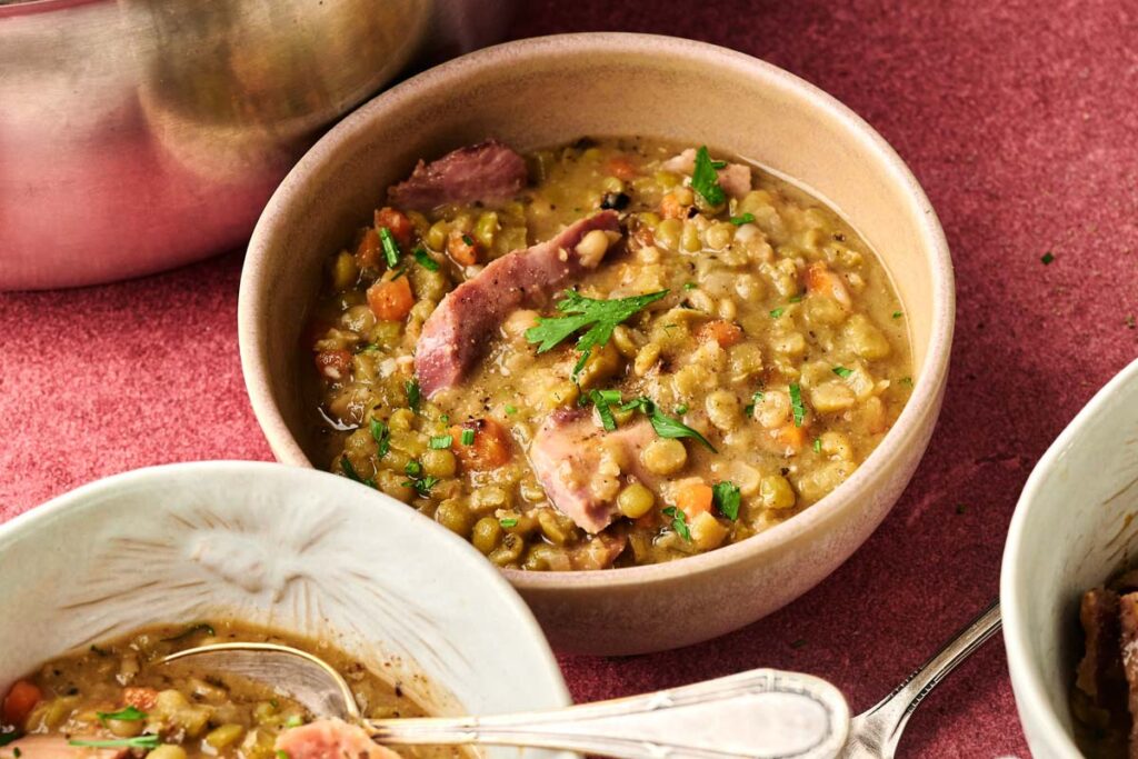 A bowl of Ham and Split Pea Soup with chunks of ham, diced carrots, and herbs is served on a red surface beside a pot and a plate with a spoon.
