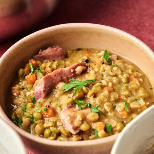A bowl of Ham and Split Pea Soup with chunks of ham, diced carrots, and fresh herbs on top, served on a vibrant red surface.