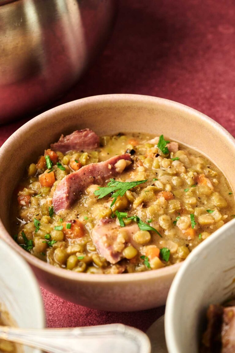 A bowl of Ham and Split Pea Soup with chunks of ham, diced carrots, and fresh herbs on top, served on a vibrant red surface.