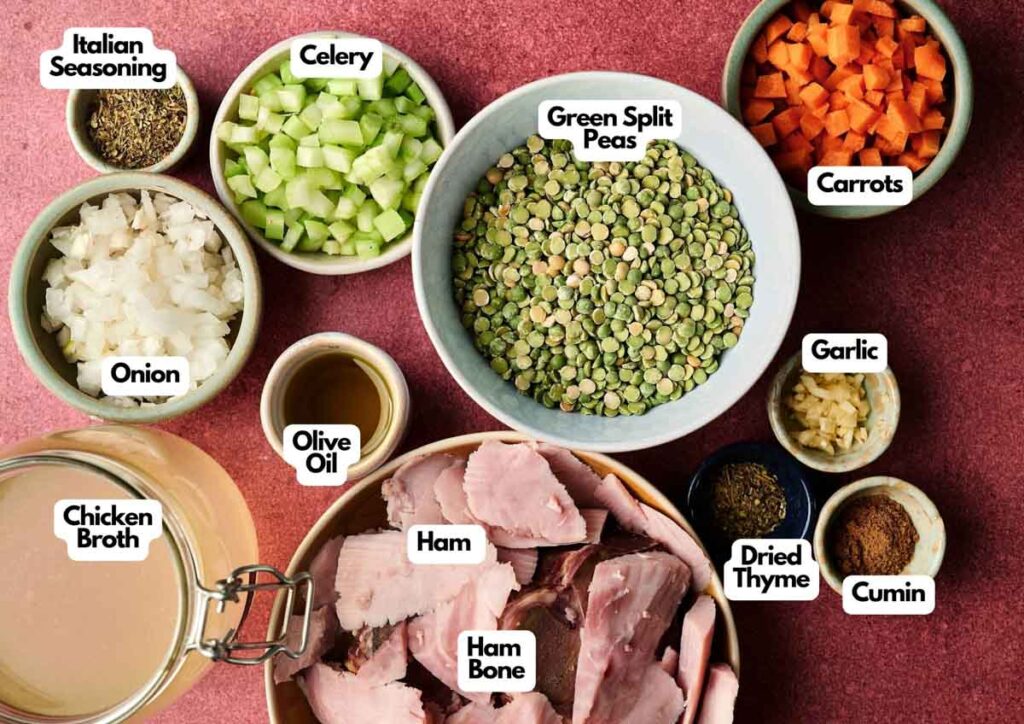 Assorted ingredients for Ham and Split Pea Soup, including green split peas, chopped vegetables, spices, ham, ham bone, chicken broth, and olive oil, arranged on a red surface.