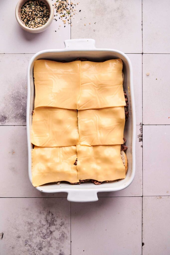 A white baking dish filled with layered ingredients for Hamburger Sliders, topped with six slices of processed cheese; seasoning in a small bowl nearby.