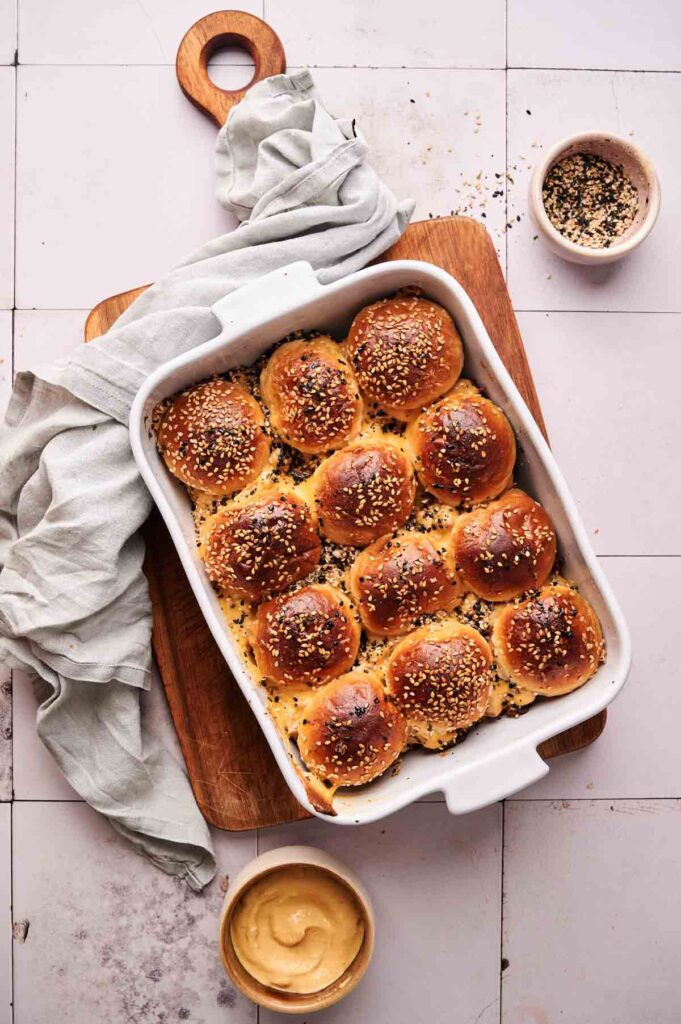 A white baking dish filled with twelve golden-brown, sesame-seeded bread rolls&mdash;perfect for Hamburger Sliders&mdash;sits on a wooden board, surrounded by small bowls of dip and seasoning.