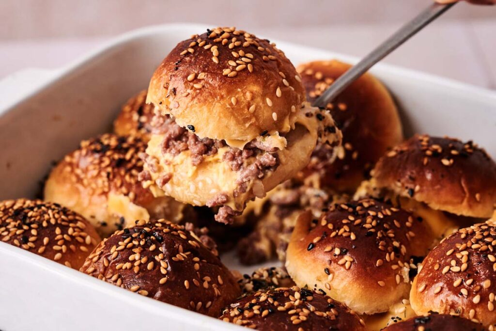 A spatula lifts a sesame-seeded hamburger slider bun filled with melted cheese and ground beef from a baking dish containing several similar sliders.