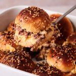 A spatula lifts a sesame-seeded hamburger slider bun filled with melted cheese and ground beef from a baking dish containing several similar sliders.