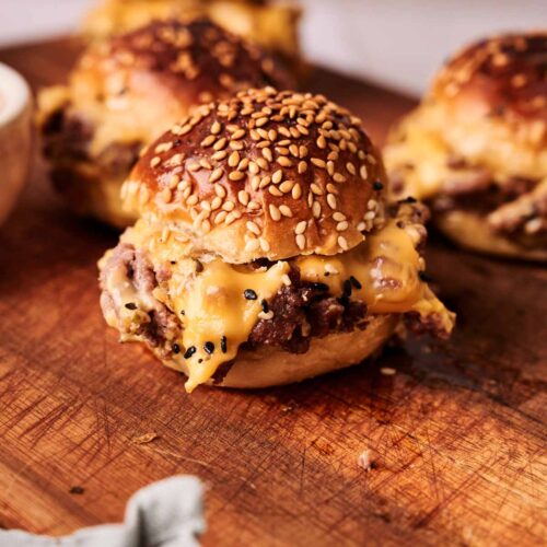 A close-up of hamburger sliders with sesame seed buns on a wooden cutting board, with melted cheese spilling out of the sides.