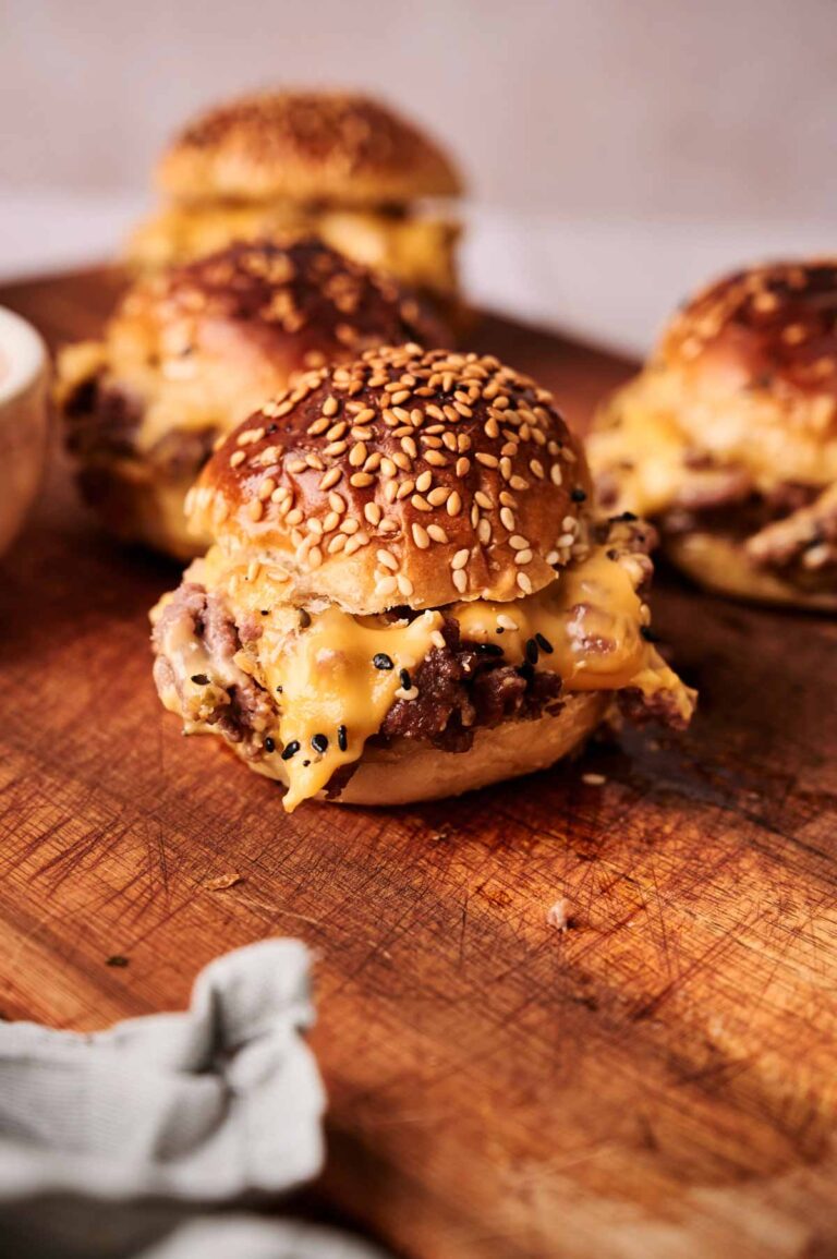 A close-up of hamburger sliders with sesame seed buns on a wooden cutting board, with melted cheese spilling out of the sides.