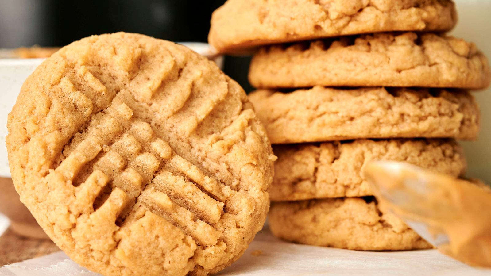 A close-up of several peanut butter cookies, with one cookie in front showing a crisscross fork pattern on top.