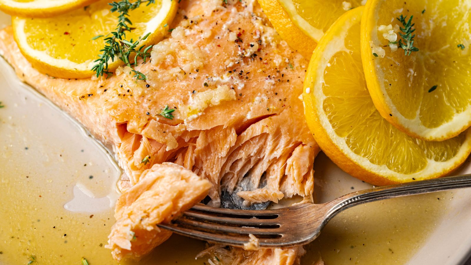 Cooked salmon fillet garnished with lemon slices and herbs, with a fork flaking the fish on a plate with sauce.