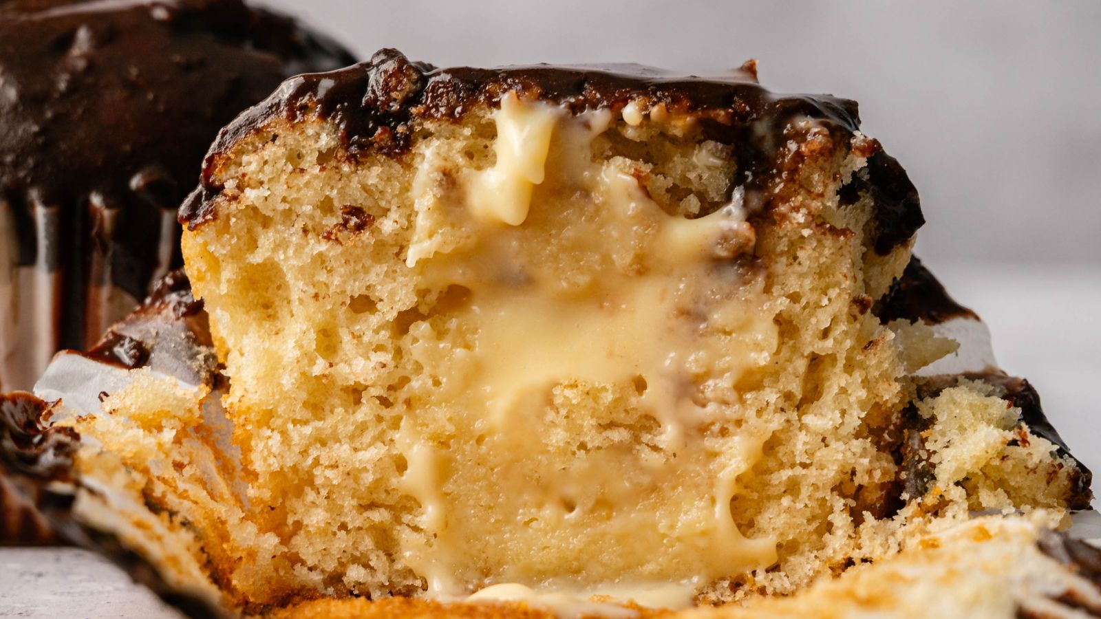 A close-up of a sliced muffin filled with creamy vanilla custard and topped with chocolate glaze.