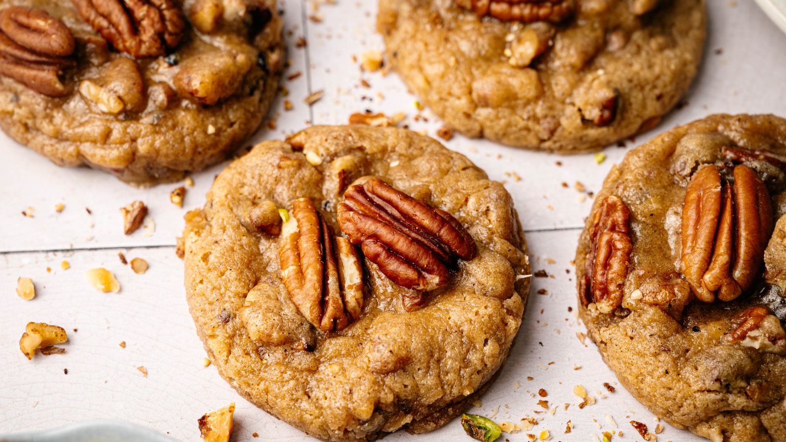Close-up of several pecan cookies on a white surface, with pecan halves on top and crumbs scattered around.