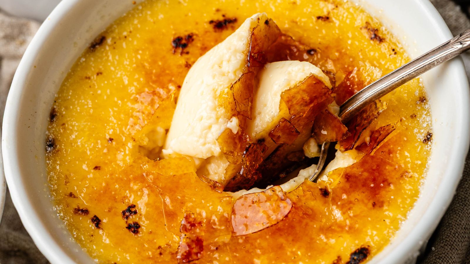 A close-up of cr&egrave;me br&ucirc;l&eacute;e in a white dish, showing a caramelized sugar crust being cracked with a spoon to reveal creamy custard underneath.