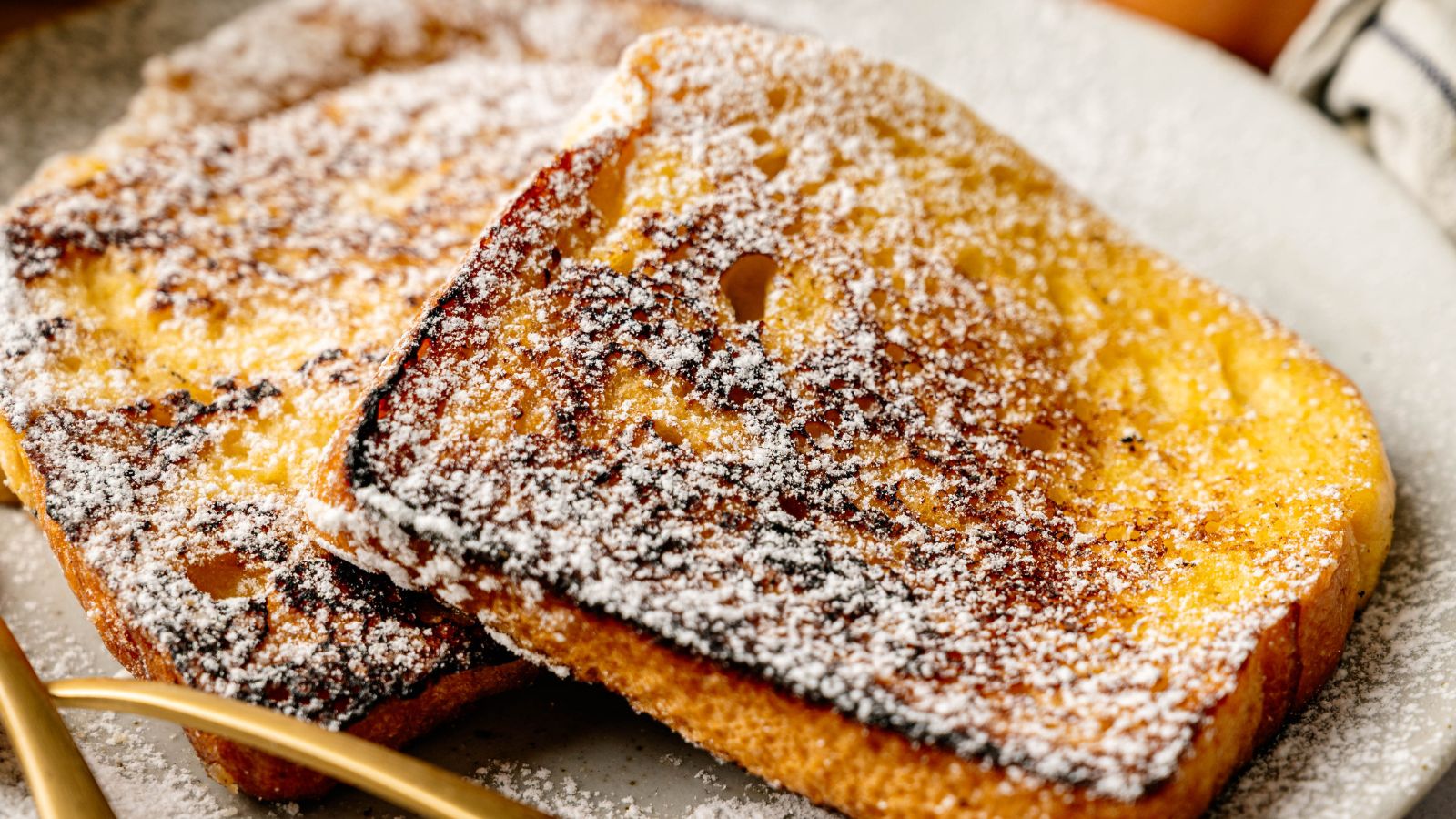 Two slices of French toast dusted with powdered sugar on a white plate, with a gold fork nearby.