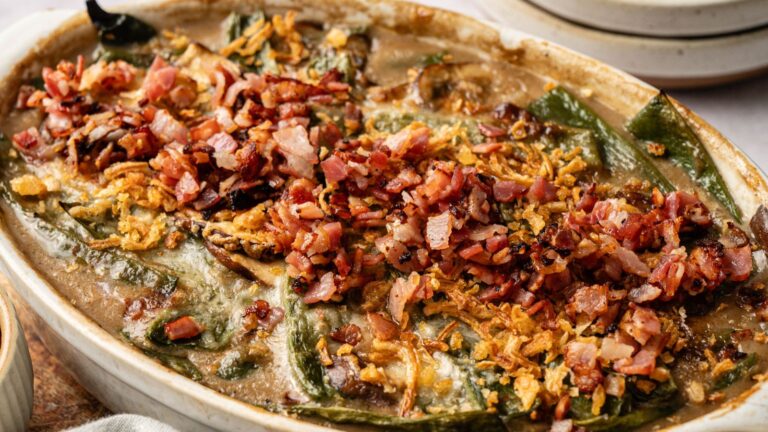A casserole dish filled with baked stuffed poblano peppers, topped with chopped bacon and crispy fried onions.