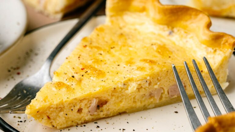 A slice of quiche on a plate with a fork beside it, showing a golden, flaky crust and a creamy, yellow filling with visible bits of ham.
