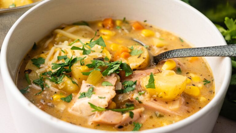 A bowl of creamy chicken and vegetable stew with potatoes, corn, carrots, and herbs, garnished with chopped parsley and shredded cheese, with a spoon in the bowl.