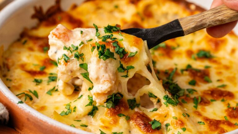 Close-up of a baked cheesy pasta dish being served with a spoon, topped with melted cheese and chopped herbs.