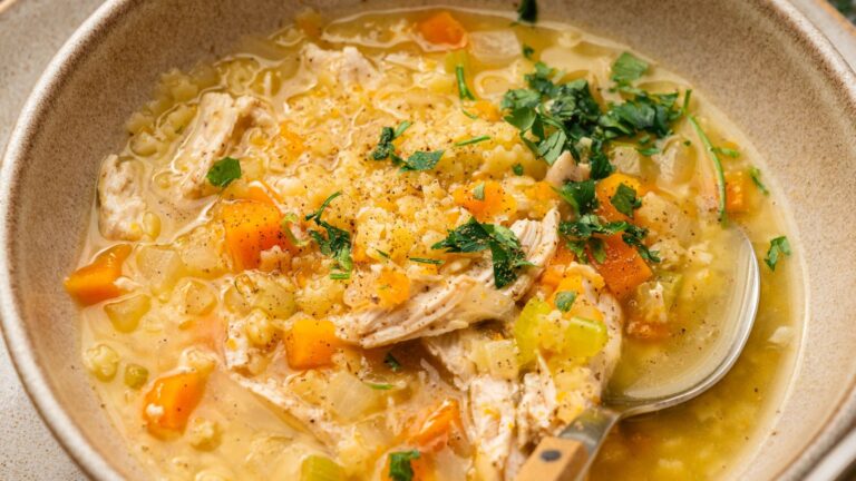 A bowl of chicken and rice soup with diced carrots, celery, onions, and fresh herbs, served with a spoon.