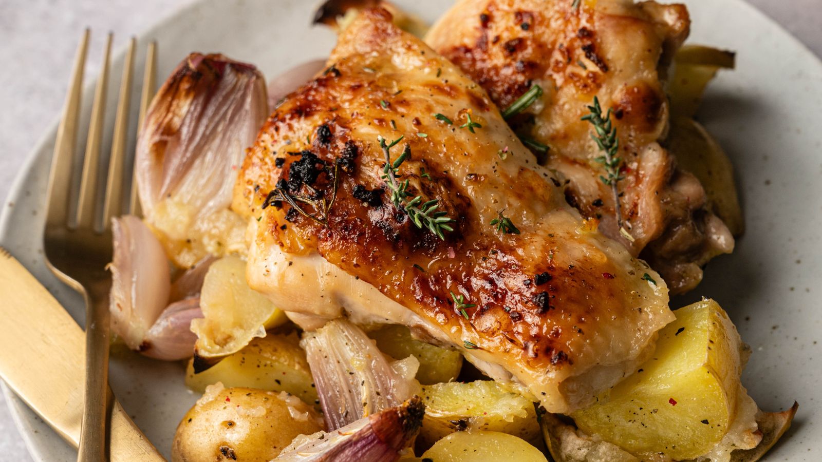 Roast chicken thighs with crispy skin served on a plate with roasted potatoes, onions, and fresh herbs, next to a gold-colored fork and knife.