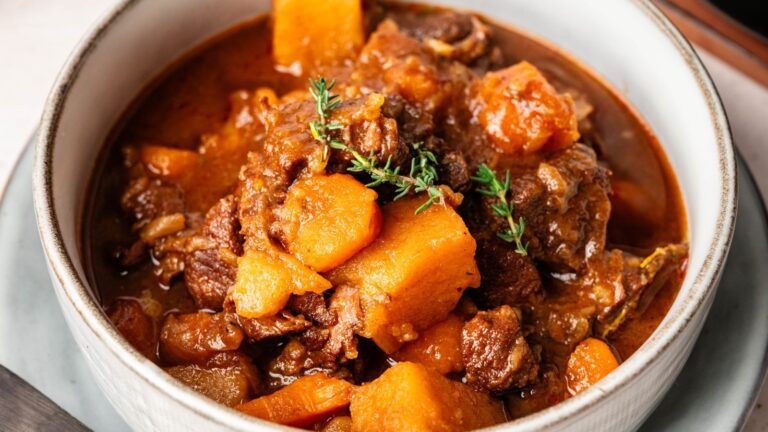 A bowl of beef stew with chunks of beef, carrots, potatoes, and a sprig of thyme in a thick, brown broth.