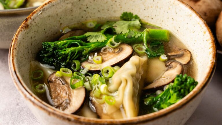 A bowl of soup with sliced mushrooms, broccoli, dumplings, chopped green onions, and fresh cilantro in a light broth.