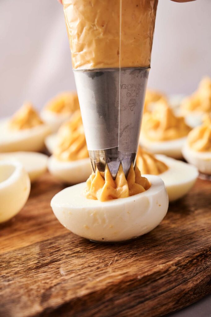 A piping bag fills a halved hard-boiled egg with creamy yolk mixture to make Million Dollar Deviled Eggs, beautifully arranged on a wooden board.