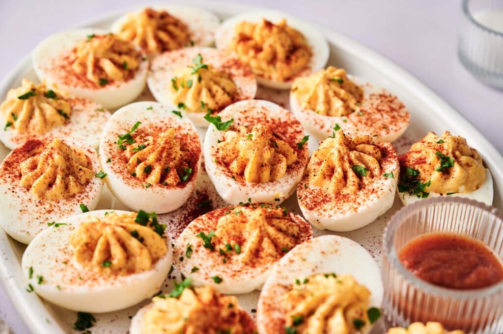 A plate of Million Dollar Deviled Eggs garnished with paprika and chopped herbs, served with a small dish of dipping sauce on the side.