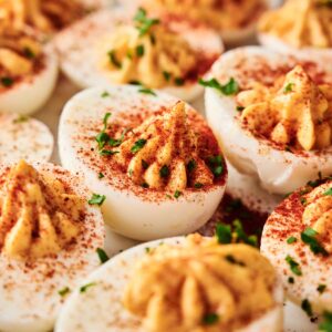 A close-up of Million Dollar Deviled Eggs topped with paprika and chopped herbs, arranged on a white serving platter.
