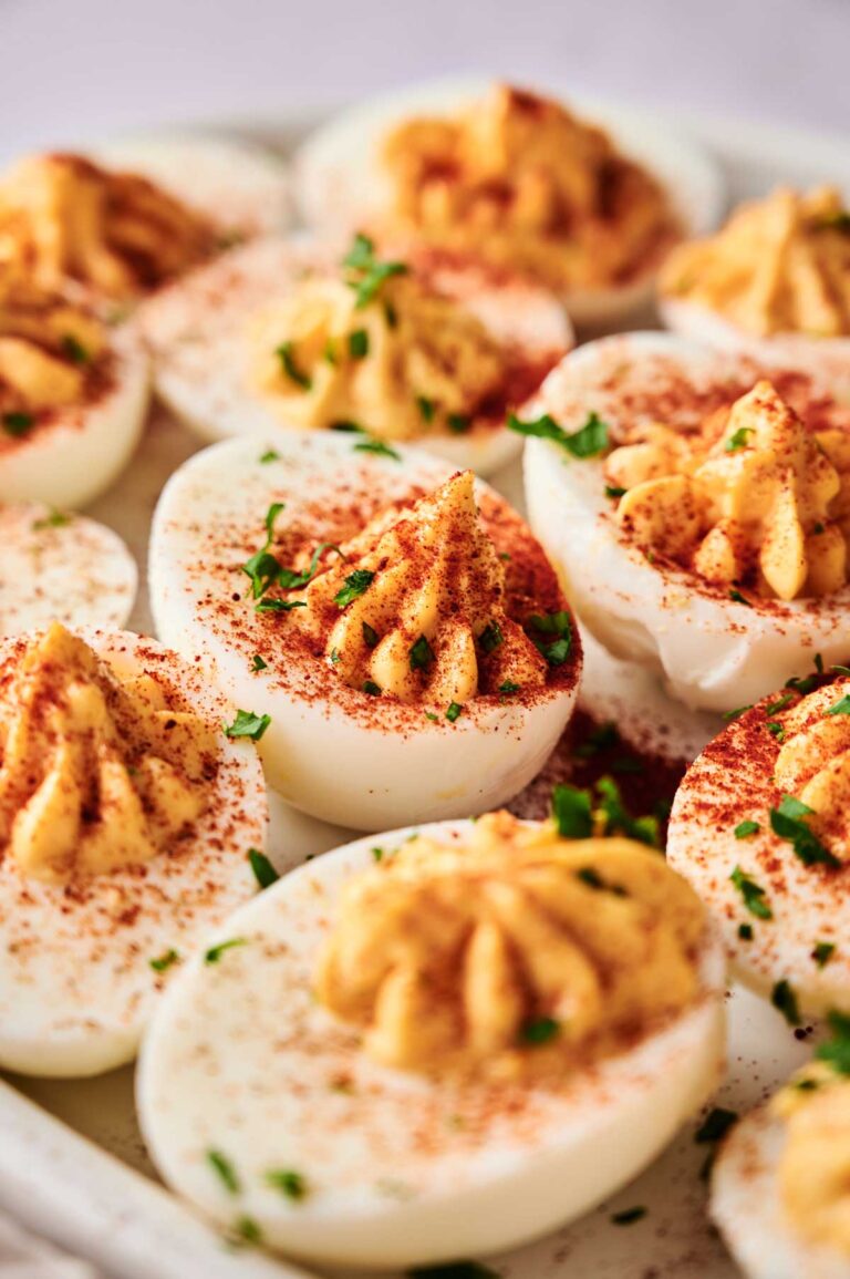 A close-up of Million Dollar Deviled Eggs topped with paprika and chopped herbs, arranged on a white serving platter.
