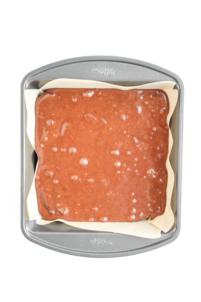 A square baking pan lined with parchment paper filled with rich, unbaked Pecan Pie Brownie batter.