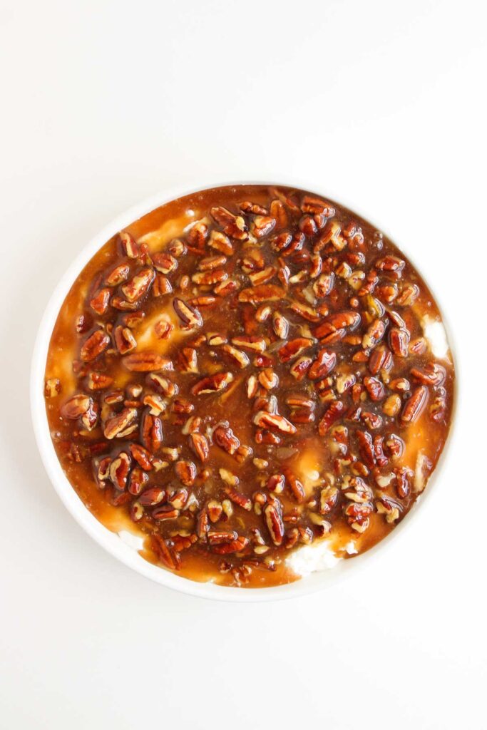 A round dish filled with creamy Pecan Pie Dip, topped with a glossy pecan and caramel sauce, photographed from above on a white background.